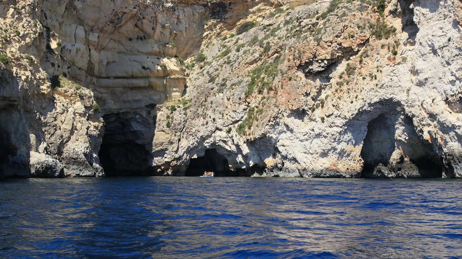 The sea caves and deep blue water of the Blue Grotto at Wied iż-Żurrieq, with a small boat below the cliffs
