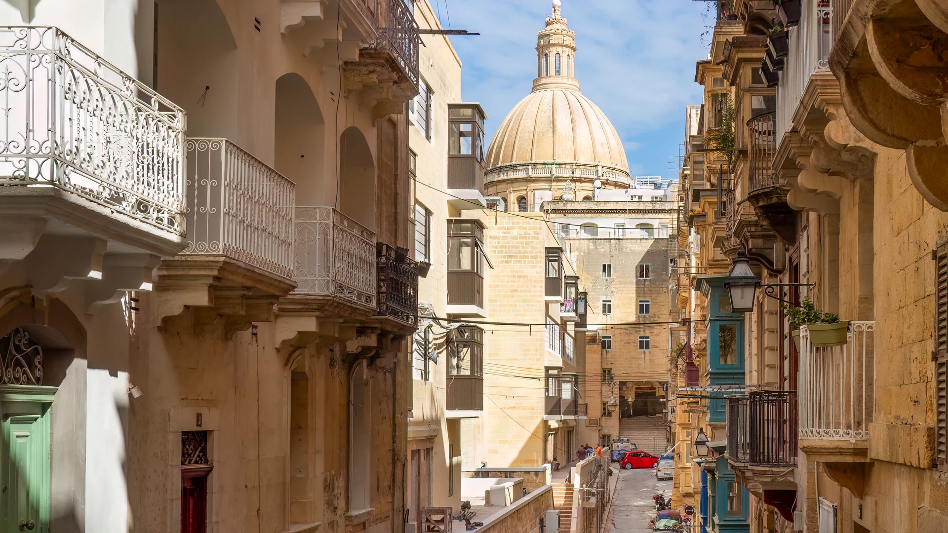 A sunlit Valletta street framing the Carmelite Church dome, with traditional Maltese balconies