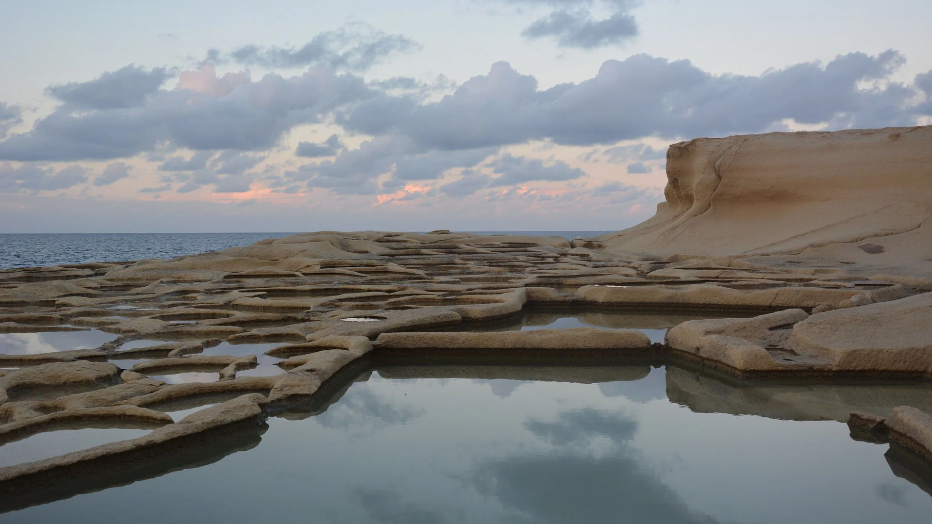 The chequerboard Xwejni salt pans on the Gozo coast at dusk, with the sea beyond