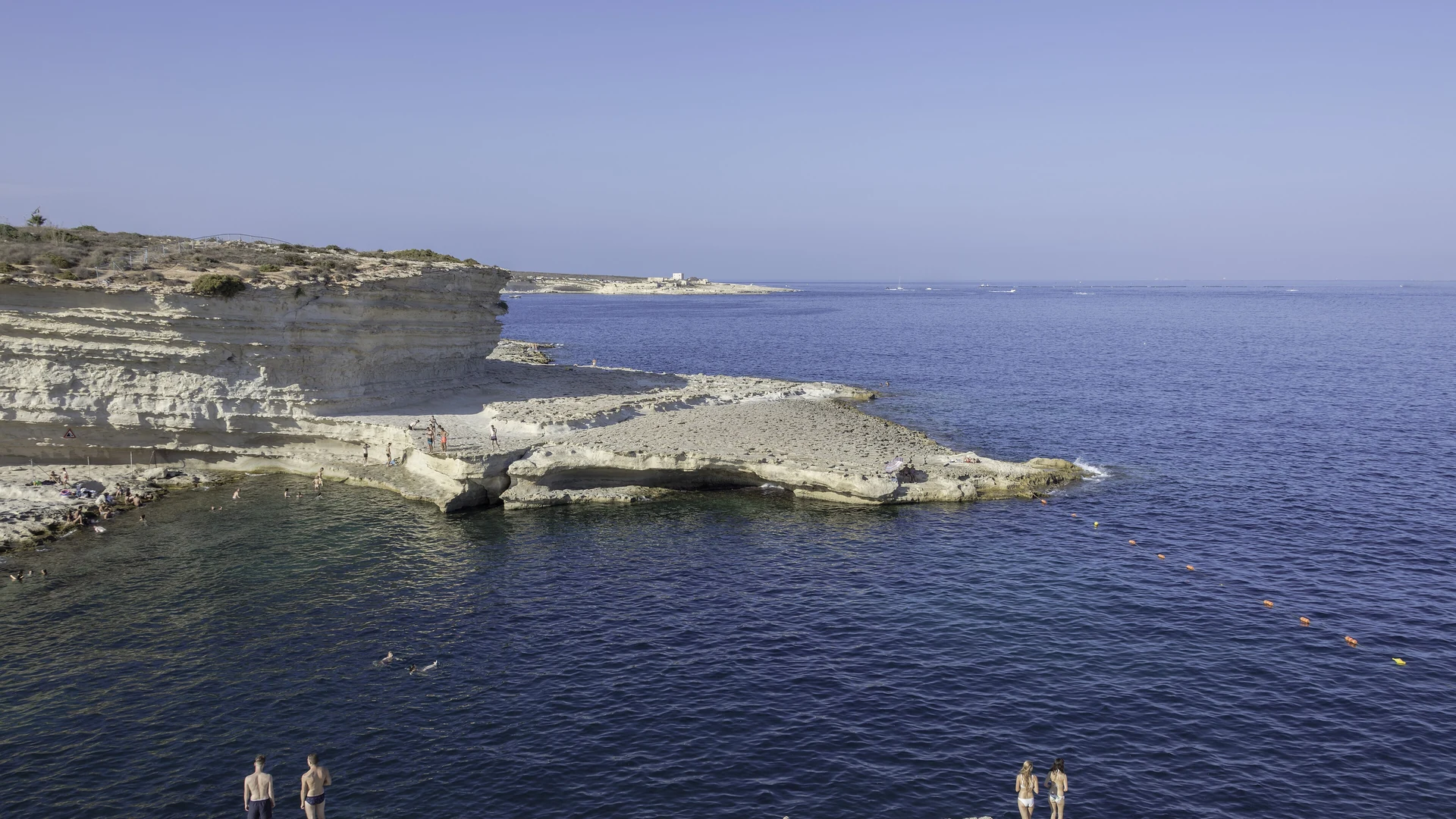 Swimmers at the rocky St Peter's Pool near Marsaxlokk, with deep blue sea under a cloudless sky