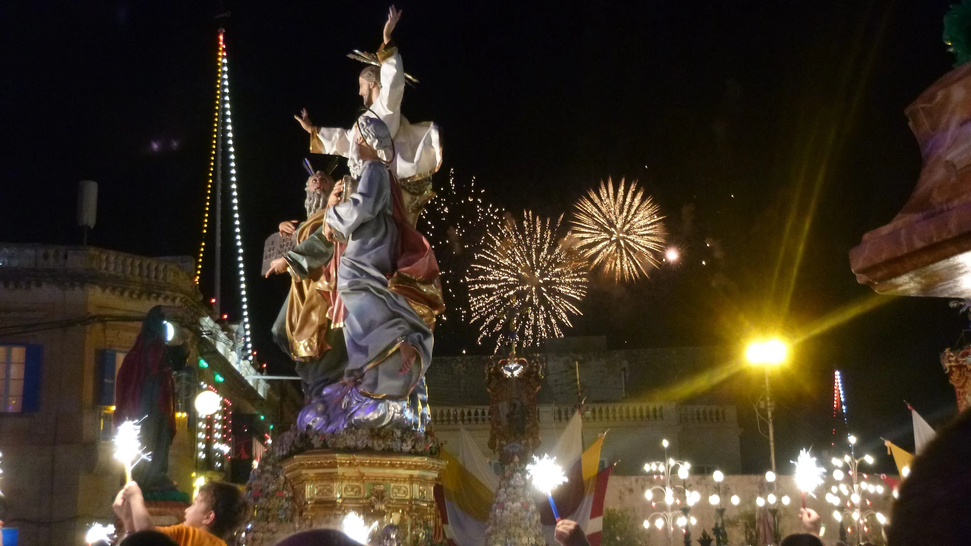 A village festa at night in Malta, with a decorated statue, fireworks and a crowd holding sparklers