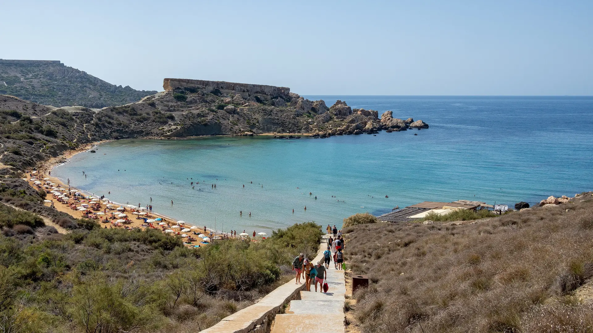 Swimmers in the turquoise water of Għajn Tuffieħa Bay, Malta, under a clear late-summer sky